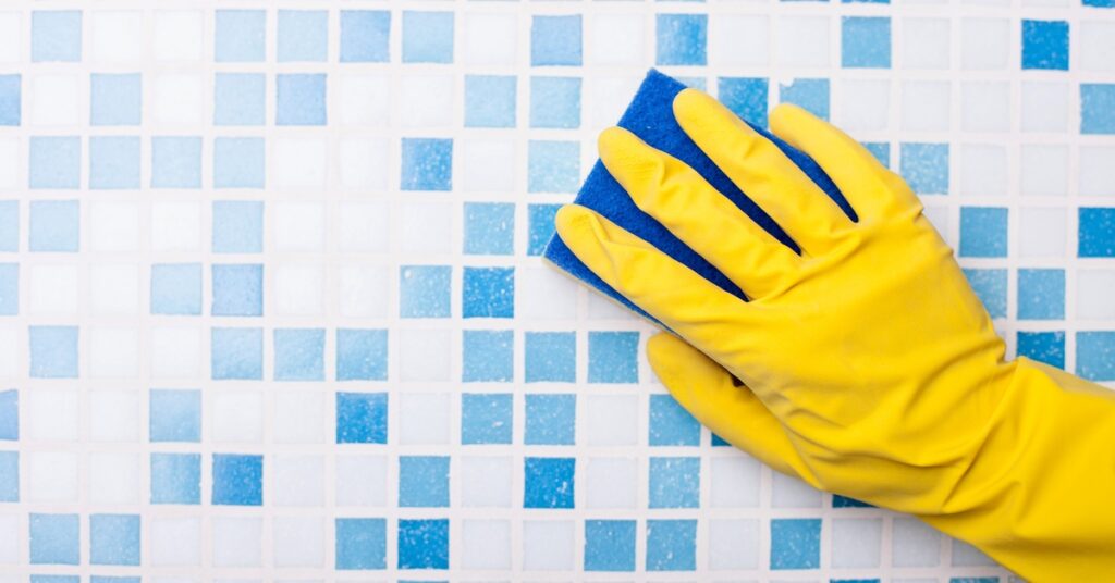 A person’s hand, wearing a yellow rubber glove, goes over a blue-and-white tiled wall with a blue sponge.