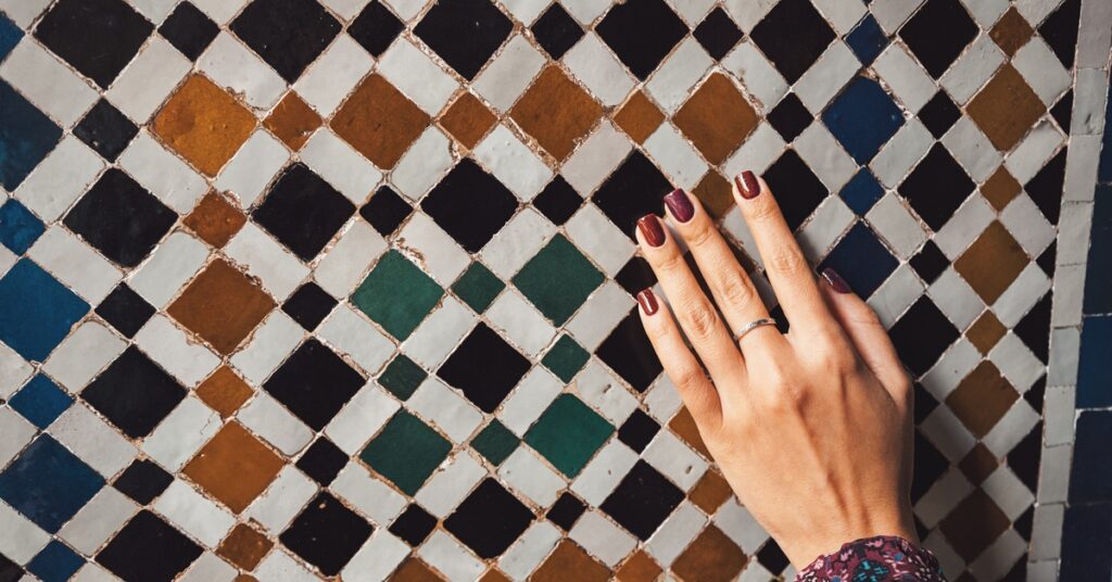 A person’s manicured hand touches a wall that’s been tiled with Moroccan Zellige tiles. The grout shows signs of age.