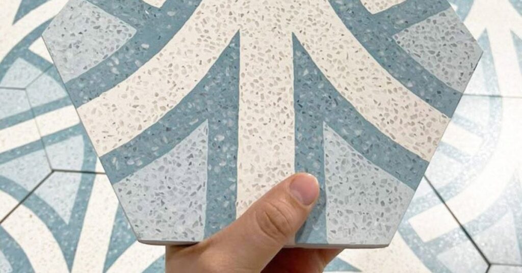A hand holding a hexagonal patterned cement tile over a matching installed floor.