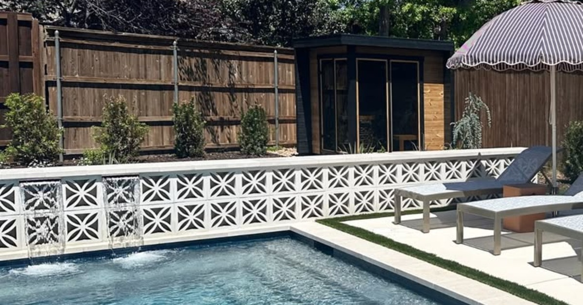 An outside environment with a pool, a lounge chair, and an umbrella. There is a wall of breeze blocks in the background.