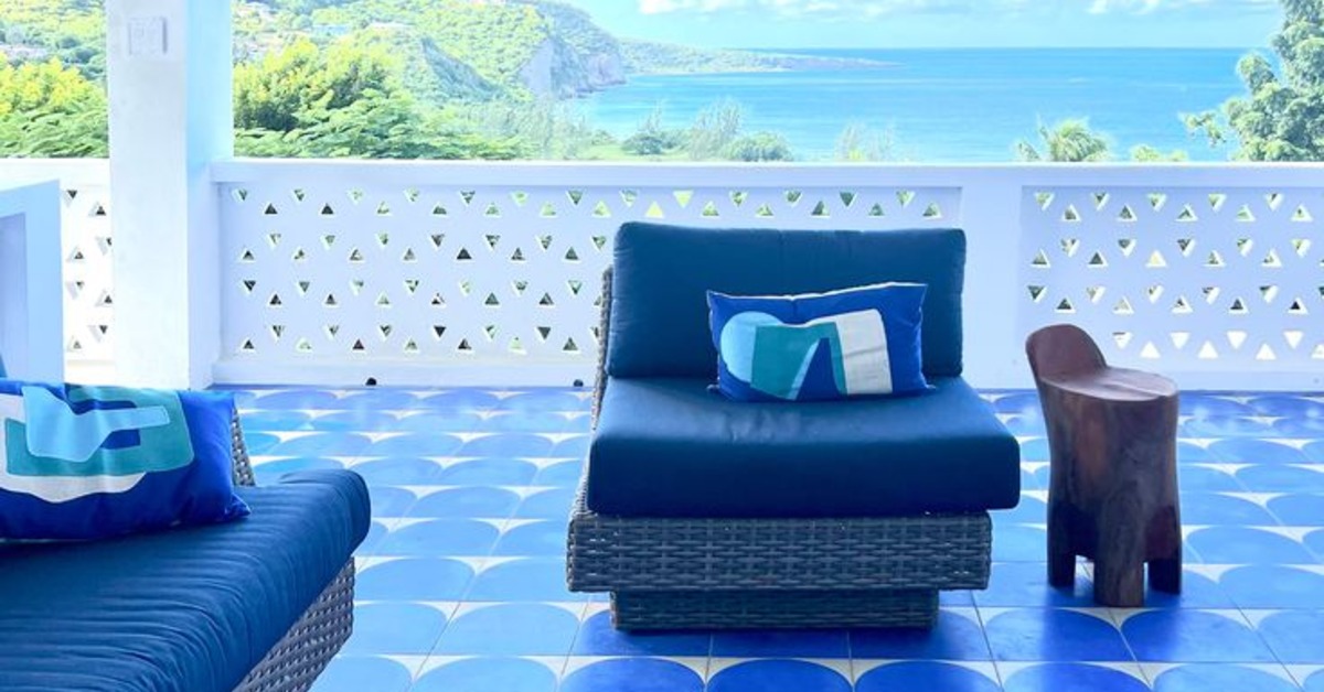 A blue chair is placed on a patio that has blue and white tiles. There is a white balcony in the background overlooking a body of water.